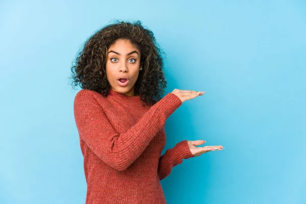 Young african american curly hair woman shocked and amazed holding a copy space between hands.