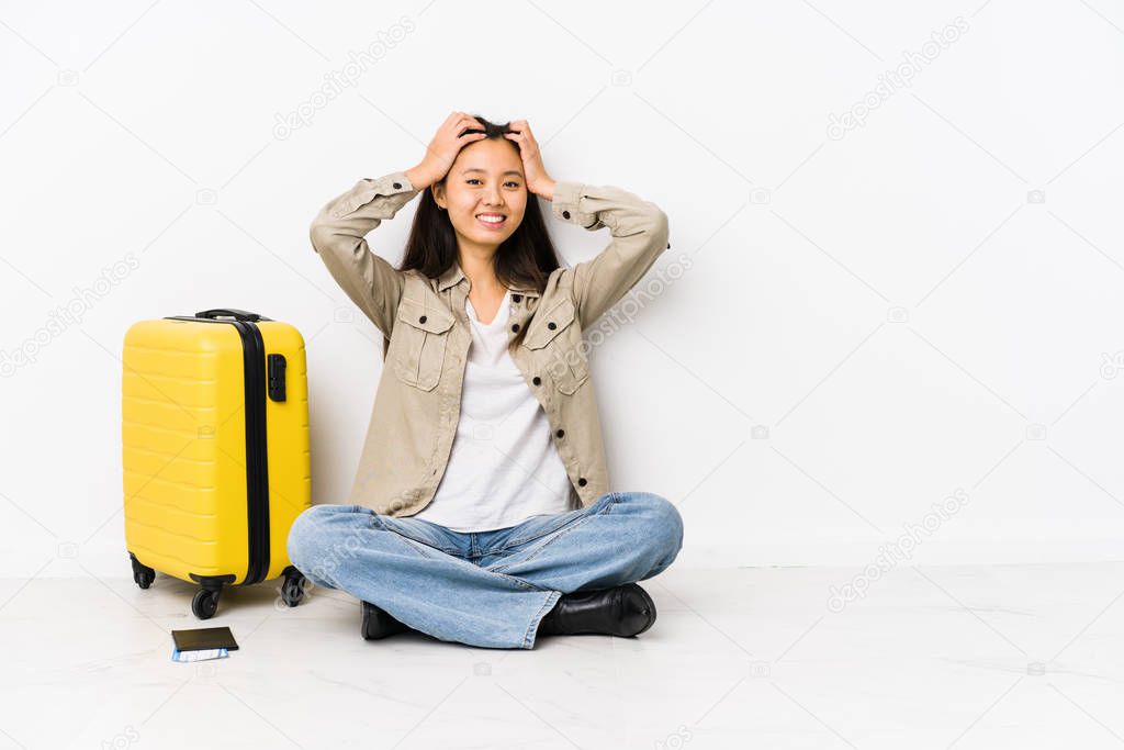 Young chinese traveler woman sitting holding a boarding passes laughs joyfully keeping hands on head. Happiness concept.