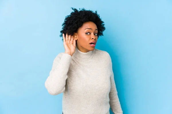 Middle aged african american woman against a blue background isolated trying to listening a gossip.
