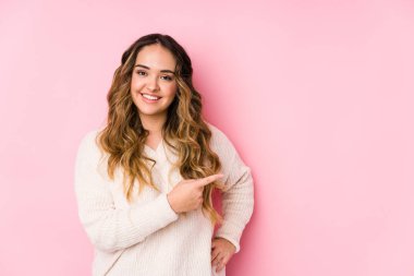Pembe arka planda poz veren kıvrımlı genç bir kadın izole edilmiş, gülümseyen ve işaret eden boş alanda bir şey gösteriyor..