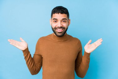 Mavi arka planı olan genç bir latin adam kollarıyla ölçek yapar, mutlu ve kendinden emin hisseder..