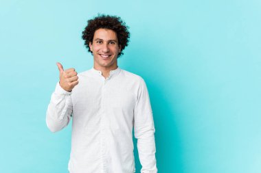 Genç, kıvırcık, olgun, zarif bir gömlek giyen, gülümseyen ve başparmağını kaldıran bir adam.