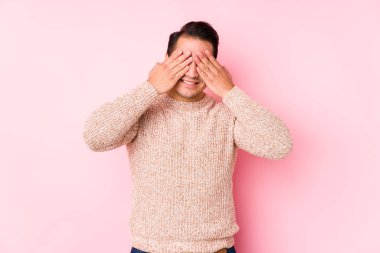 Young curvy man posing in a pink background isolated covers eyes with hands, smiles broadly waiting for a surprise.
