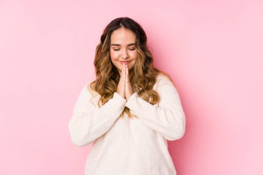 Pembe arka planda poz veren kıvrımlı genç bir kadın. El ele tutuşup dua ediyor. Kendine güveniyor..