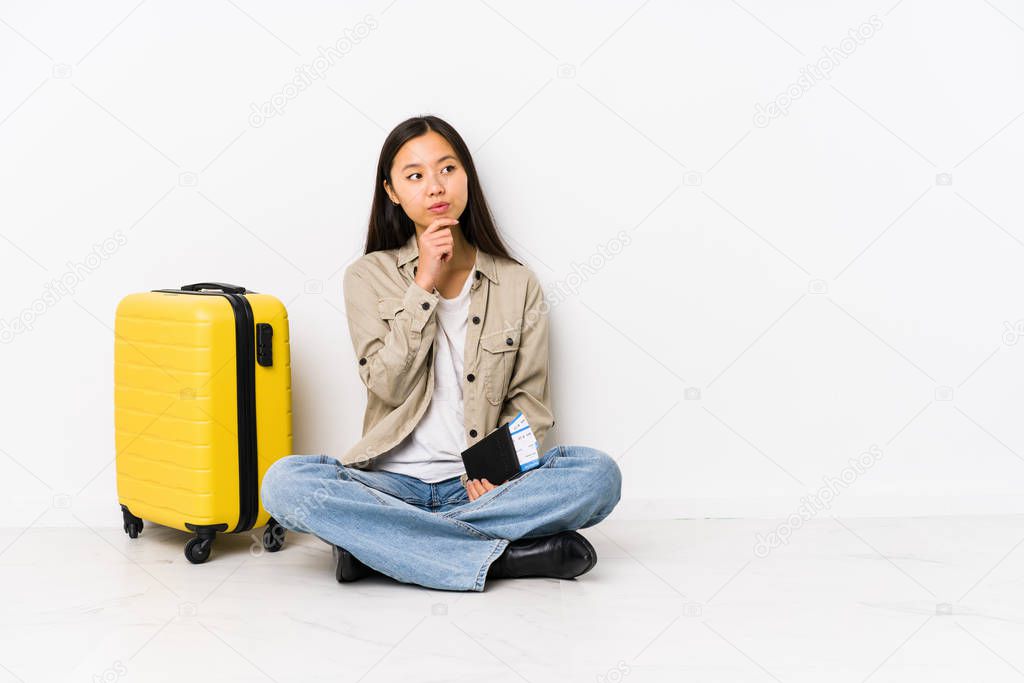 Young chinese traveler woman sitting holding a boarding passes looking sideways with doubtful and skeptical expression.