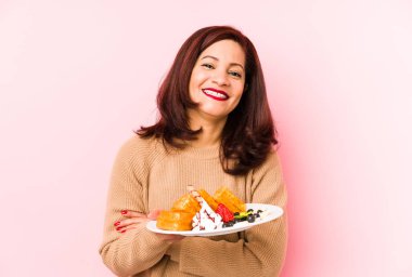 Orta yaşlı Latin bir kadın elinde waffle tutarak gülüyor ve eğleniyor..