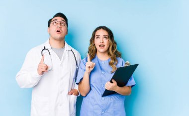 Young doctor couple posing in a blue background isolated pointing upside with opened mouth.