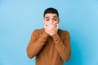 Mavi arka planı olan genç bir latin adam elleri ile ağzını kapatırken şok oldu..