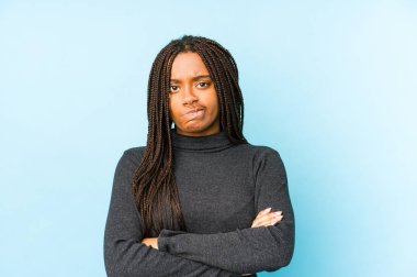 Mavi arka planda izole edilmiş, hoşnutsuzca kaşlarını çatmış, kollarını kavuşturmuş genç Afrikalı Amerikalı kadın..