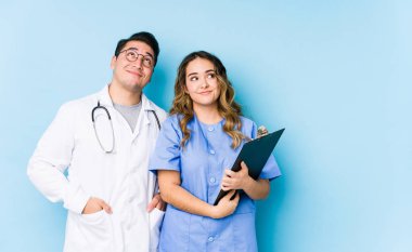 Young doctor couple posing in a blue background isolated dreaming of achieving goals and purposes