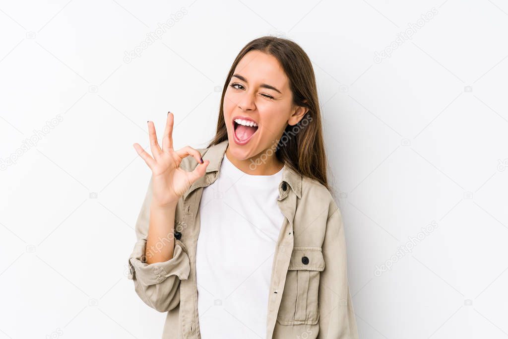 Young caucasian woman  isolated winks an eye and holds an okay gesture with hand.