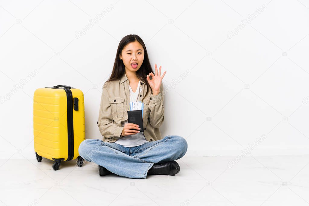 Young chinese traveler woman sitting holding a boarding passes winks an eye and holds an okay gesture with hand.