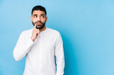 Mavi arka planı olan genç bir latin adam şüpheli ve şüpheci bir ifadeyle yanlamasına bakıyor..