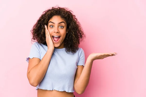 Pembe arka planı olan genç Afrikalı Amerikalı bir kadın avuç içinde fotokopi alanı tutuyor, el üstünde tutuyor. Şaşırmış ve mutlu..