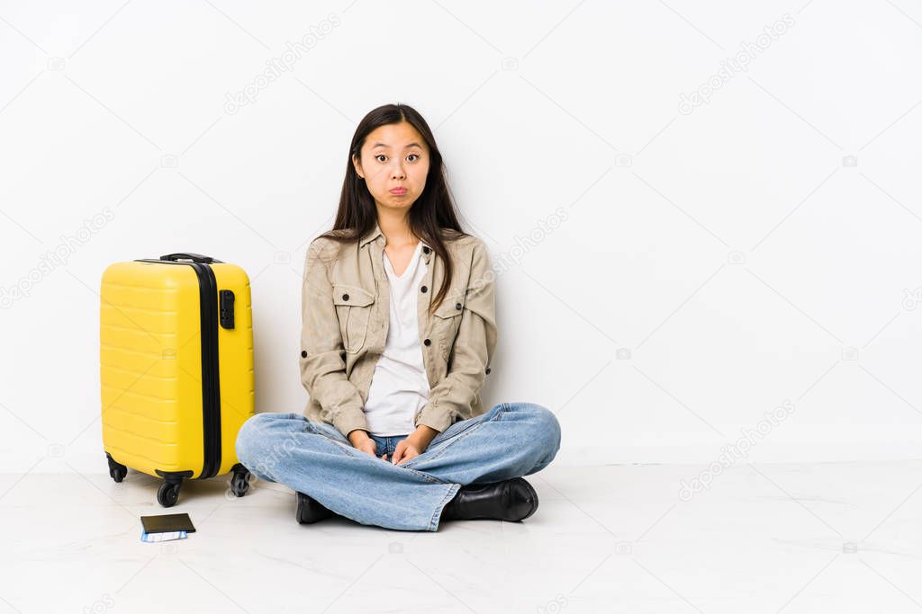 Young chinese traveler woman sitting holding a boarding passes shrugs shoulders and open eyes confused.