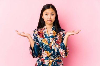 Young chinese woman wearing a kimono pajama isolated confused and doubtful shrugging shoulders to hold a copy space.
