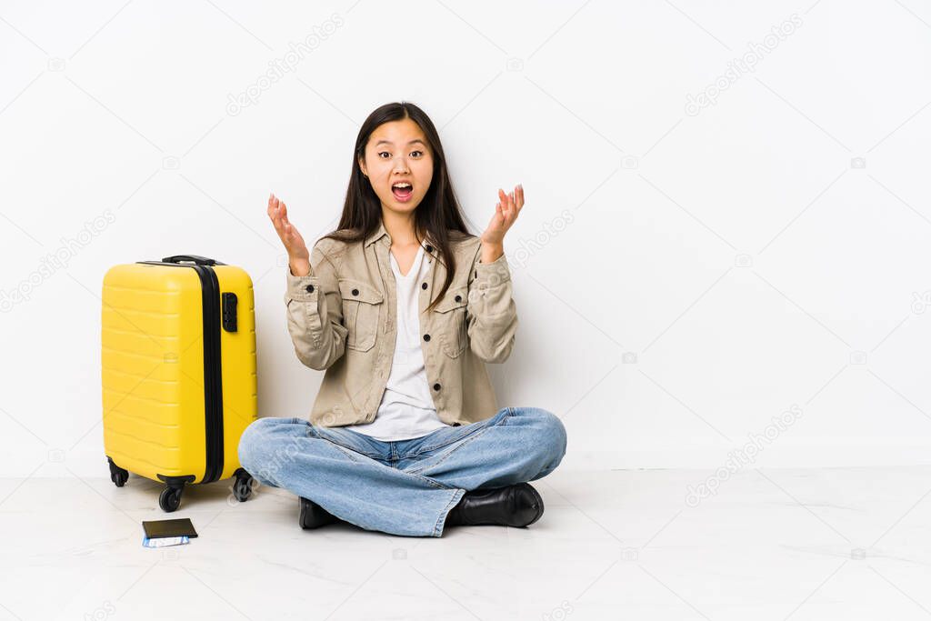 Young chinese traveler woman sitting holding a boarding passes receiving a pleasant surprise, excited and raising hands.