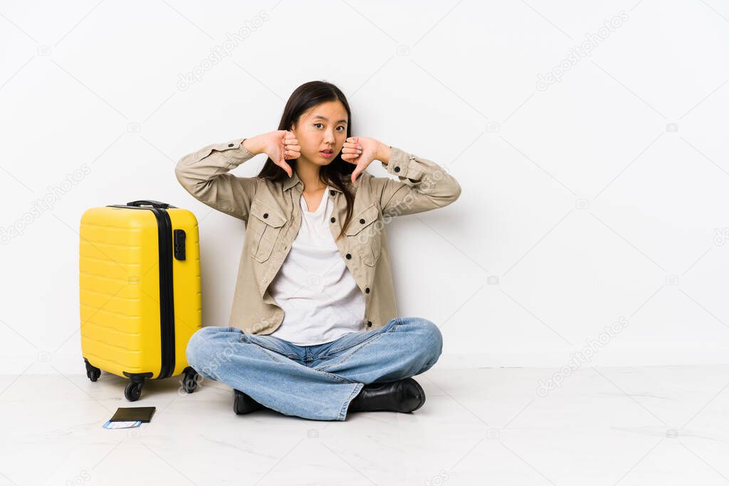 Young chinese traveler woman sitting holding a boarding passes showing thumb down and expressing dislike.