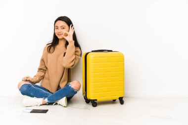 Genç Çinli gezgin kadın elinde bir bavulla yerde oturuyor neşeli ve kendinden emin bir şekilde..