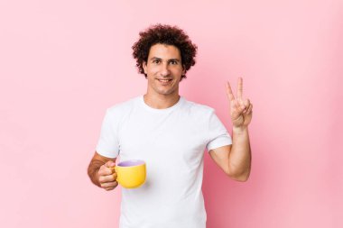 Beyaz, kıvırcık, elinde çay fincanı tutan genç adam zafer işareti gösteriyor ve gülümsüyor..