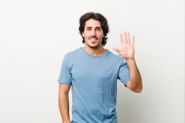 Beyaz arka planda gülümseyen genç yakışıklı adam parmaklarıyla beş numarayı gösteriyor..