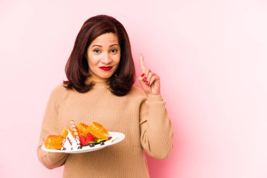 Orta yaşlı, elinde waffle tutan Latin bir kadın bir numarayı parmağıyla gösteriyor..