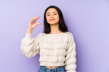 Mor bir arka planda izole edilmiş genç Çinli bir kadın işaret parmaklarıyla küçük bir şey tutuyor, gülümsüyor ve kendinden emin..