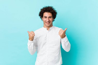 Genç, kıvırcık, olgun, zarif bir gömlek giyen, iki parmağını da kaldıran, gülümseyen ve kendinden emin bir adam..