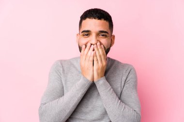 Pembe arka planda genç bir latin adam bir şeye gülüyor, elleri ile ağzını kapatıyor..