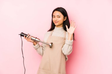 Bir mikser tutan genç Çinli kadın izole edilmiş, parmaklarıyla bir cep telefonu hareketi gösteriyor..