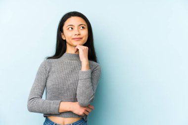 Genç Çinli bir kadın mavi bir arka planda izole edilmiş şüpheli ve şüpheci bir ifadeyle yana bakıyor..