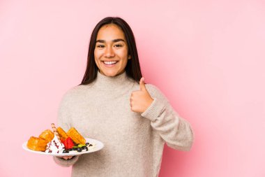 Genç Asyalı kadın bir waffle tutuyor yalnız, gülümsüyor ve başparmağını kaldırıyor.