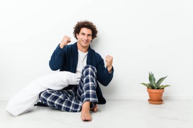 Olgun bir adam pijamasını giymiş evde oturmuş zafer kazandıktan sonra yumruğunu kaldırıyor..