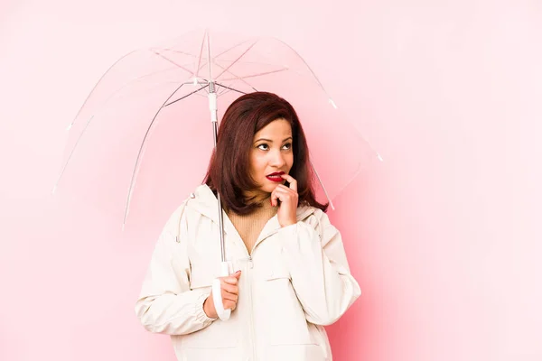 Middle age latin woman wearing an umbrella isolated relaxed thinking about something looking at a copy space.