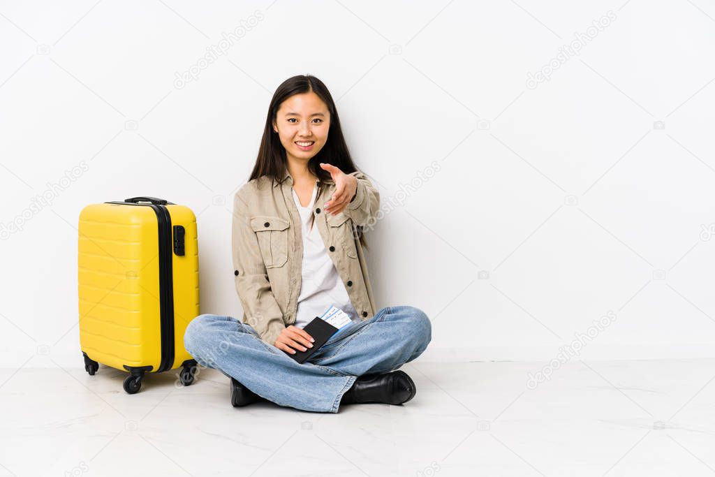 Young chinese traveler woman sitting holding a boarding passes stretching hand at camera in greeting gesture.