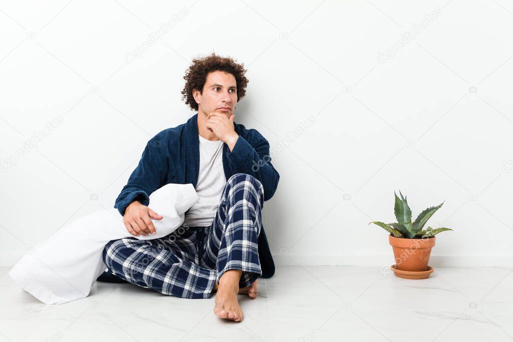 Mature man wearing pajama sitting on house floor looking sideways with doubtful and skeptical expression.