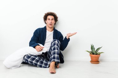 Mature man wearing pajama sitting on house floor impressed holding copy space on palm.