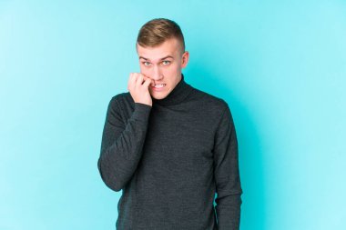 Mavi arka planda genç beyaz bir adam tırnaklarını yiyor, gergin ve çok endişeli..