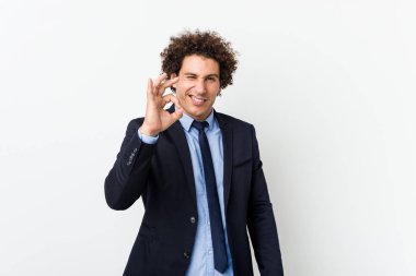 Young business curly man against white background winks an eye and holds an okay gesture with hand.