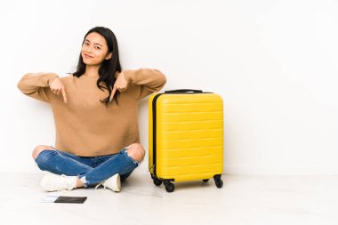 Young chinese traveler woman sittting on the floor with a suitcase isolated points down with fingers, positive feeling.