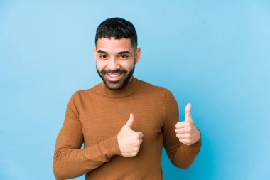 Young latin man against a blue  background isolated raising both thumbs up, smiling and confident.