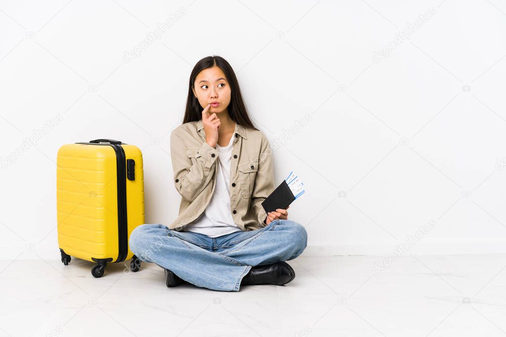 Young chinese traveler woman sitting holding a boarding passes looking sideways with doubtful and skeptical expression.