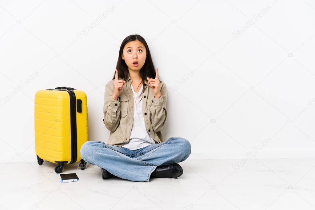 Young chinese traveler woman sitting holding a boarding passes pointing upside with opened mouth.