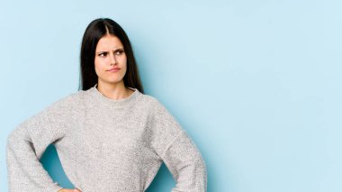 Kafkasyalı genç bir kadın mavi arka planda izole edilmiş, kafası karışmış, şüpheli ve kendinden emin değil..