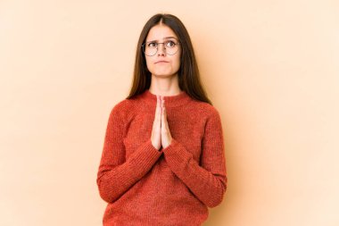 Beyaz tenli genç bir kadın, bej arka planda izole edilmiş, el ele tutuşmuş, ağız kenarında dua ediyor, kendinden emin..