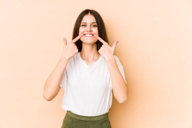 Beyazların arka planında izole edilmiş genç beyaz kadın gülümsüyor, parmaklarını ağıza doğrultuyor..