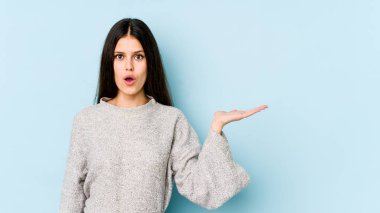 Mavi arka planda izole edilmiş genç beyaz kadın, avuç içinin fotokopi alanını tutmaktan etkilenmiş..