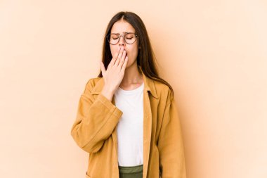 Beyaz tenli genç bir kadın bej arka planda izole edilmiş esniyor. Ağzı eliyle kapatan yorgun bir hareket sergiliyor..