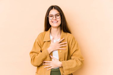 Beyaz tenli genç bir kadın bej arka planda izole edilmiş mutlu bir şekilde gülüyor ve ellerini karnının üstünde tutarak eğleniyor..