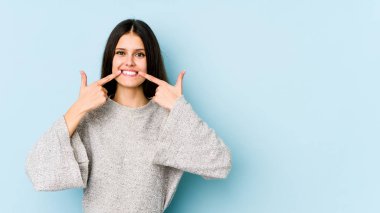 Mavi arka planda izole edilmiş genç beyaz kadın gülümsüyor, parmaklarını ağıza doğrultuyor..